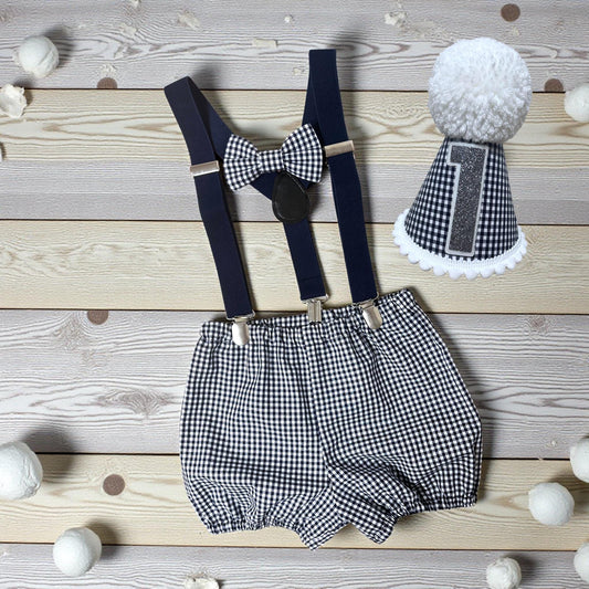 Handmade baby boy first birthday outfit with navy gingham shorts, navy suspender braces, bow tie and matching party hat featuring glitter number and pom-pom on wooden background.