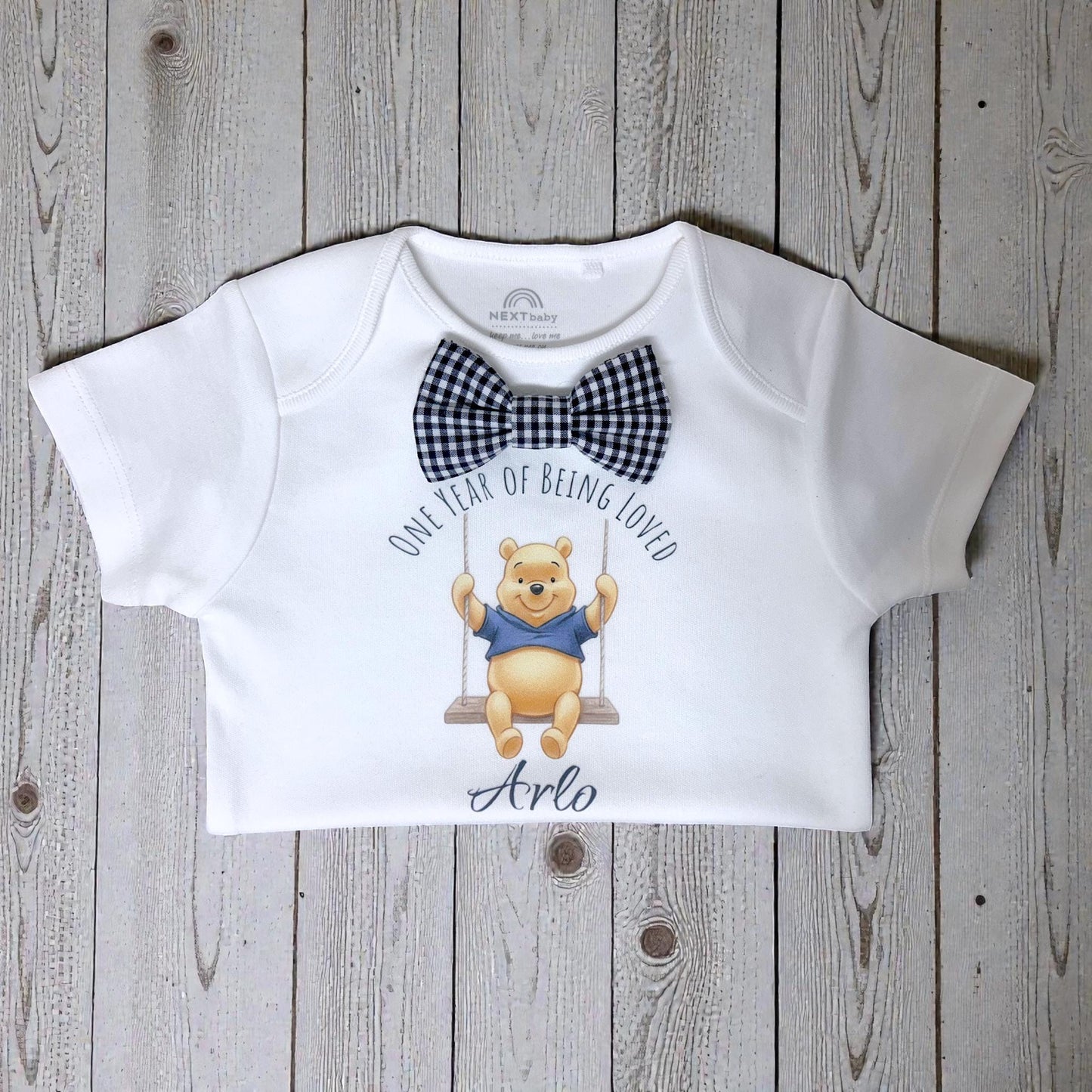 Personalised baby boy birthday T-shirt with honey bear illustration and name, displayed flat on wooden background.