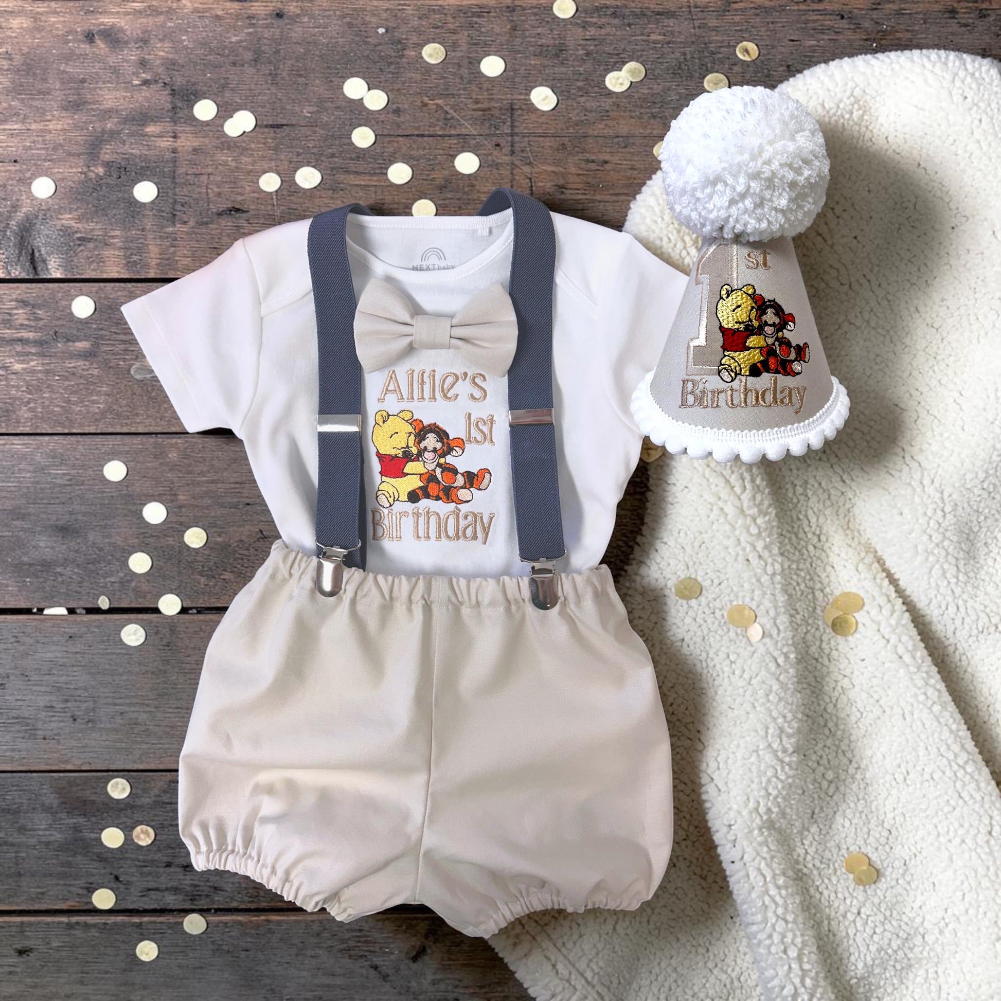 A baby outfit with a bow tie, suspenders, and a Winnie the Pooh-themed birthday cap, along with confetti on a wooden surface.