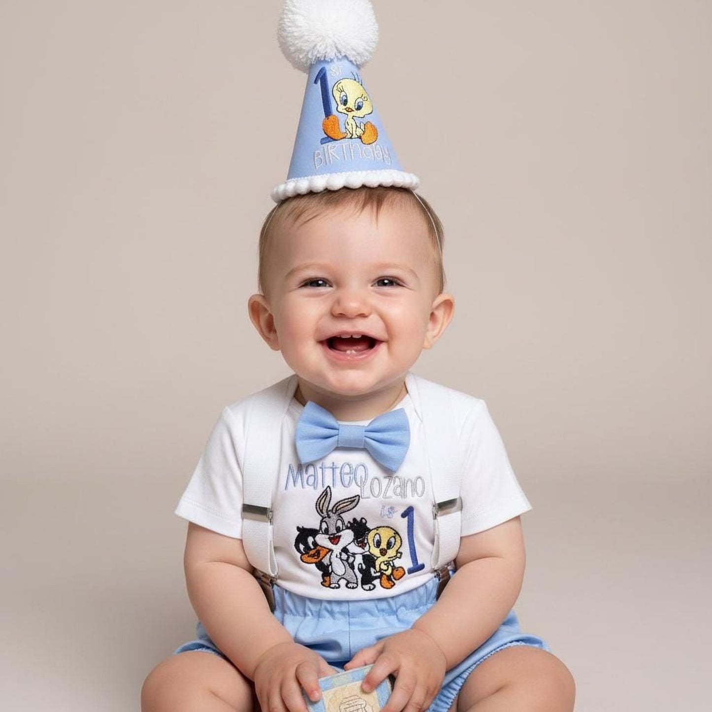 A smiling baby boy wearing a birthday hat with a cartoon character and the number 1, sitting on a plain background.