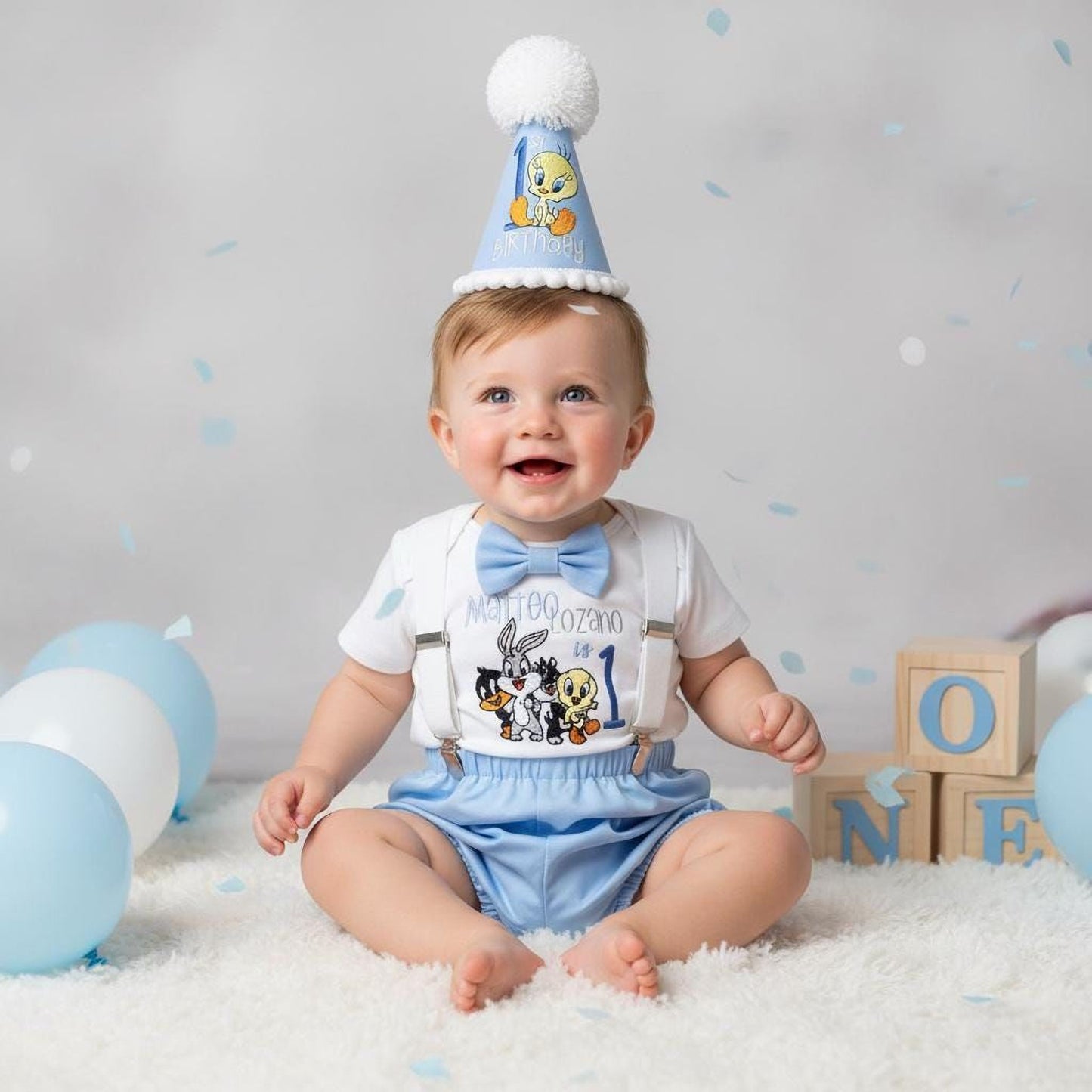 A smiling baby boy wearing a birthday hat and sitting on a white blanket surrounded by blue balloons and wooden blocks.
