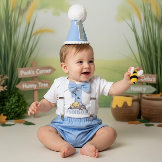 Custom Winnie the Pooh first birthday outfit with name embroidery, baby boy wearing blue bow tie and suspenders holding a bee toy.