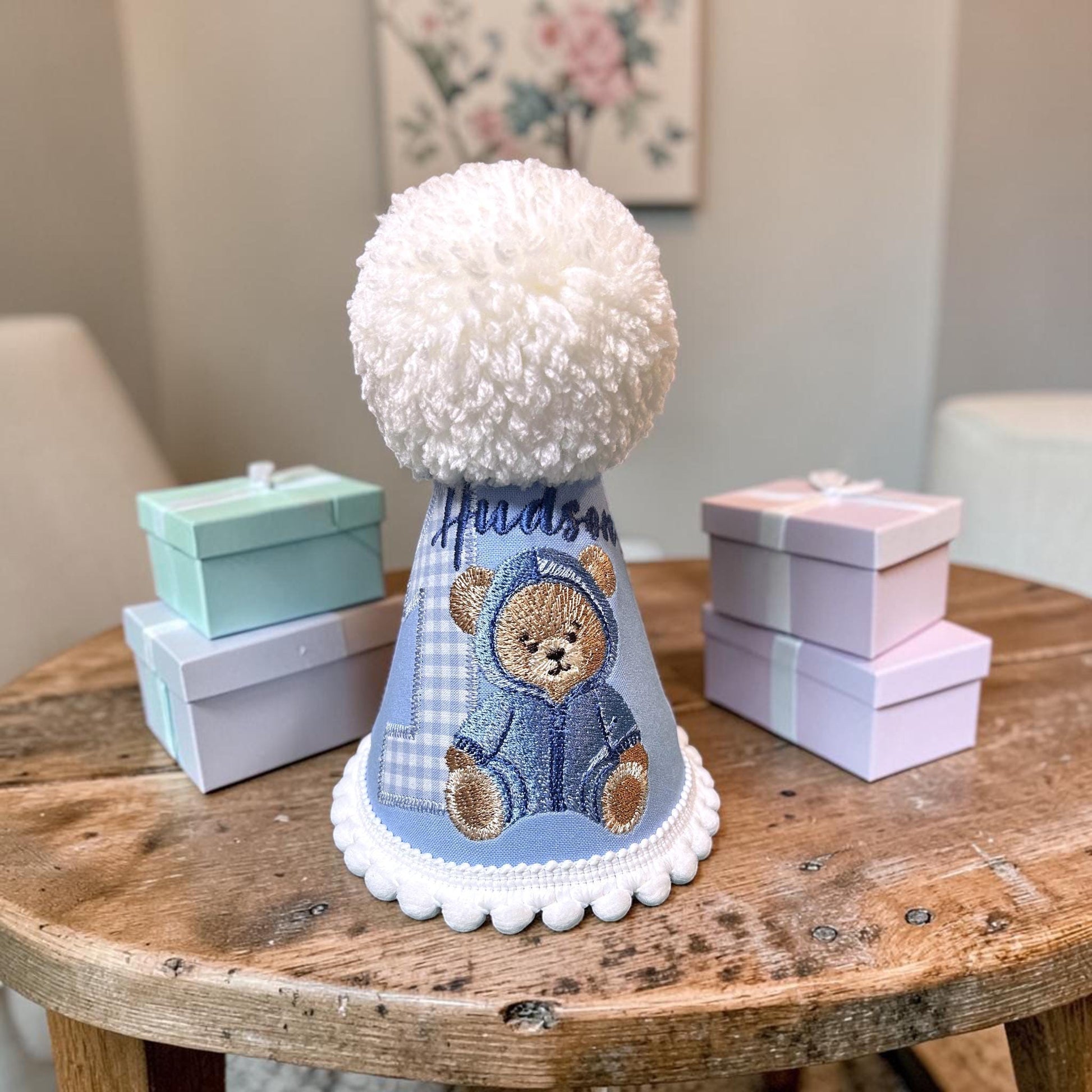 Handmade personalised blue teddy bear birthday hat with embroidered bear design, gingham number applique, white pom pom topper and decorative trim for first birthday celebrations.