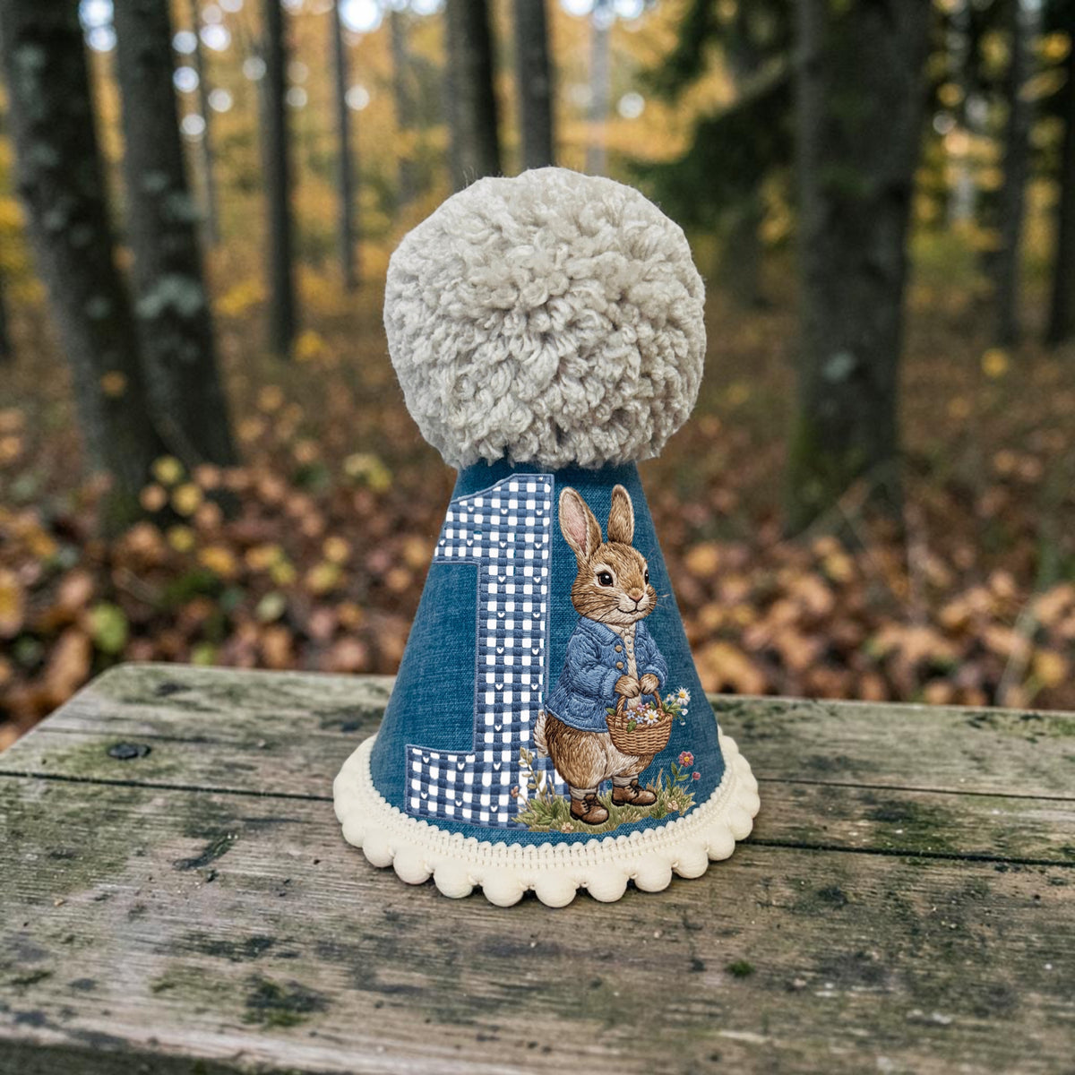 Handmade blue Peter Rabbit first birthday party hat with printed rabbit illustration, blue gingham number one, cream pom-pom trim and large fluffy cream yarn pom-pom, photographed in a woodland setting.
