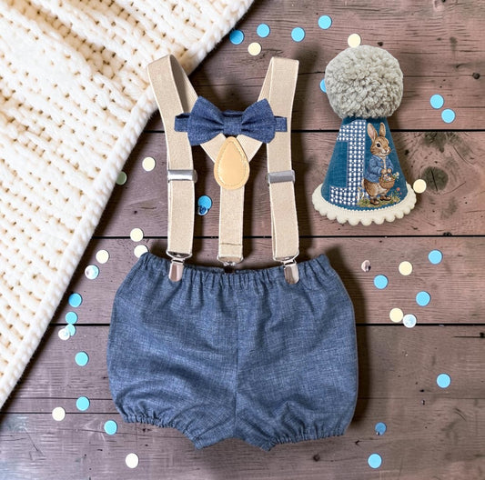 Peter Rabbit first birthday accessories set with handmade denim-blue bloomer shorts, matching blue bow, beige suspender braces and blue printed rabbit party hat with gingham number one and cream pom-pom.