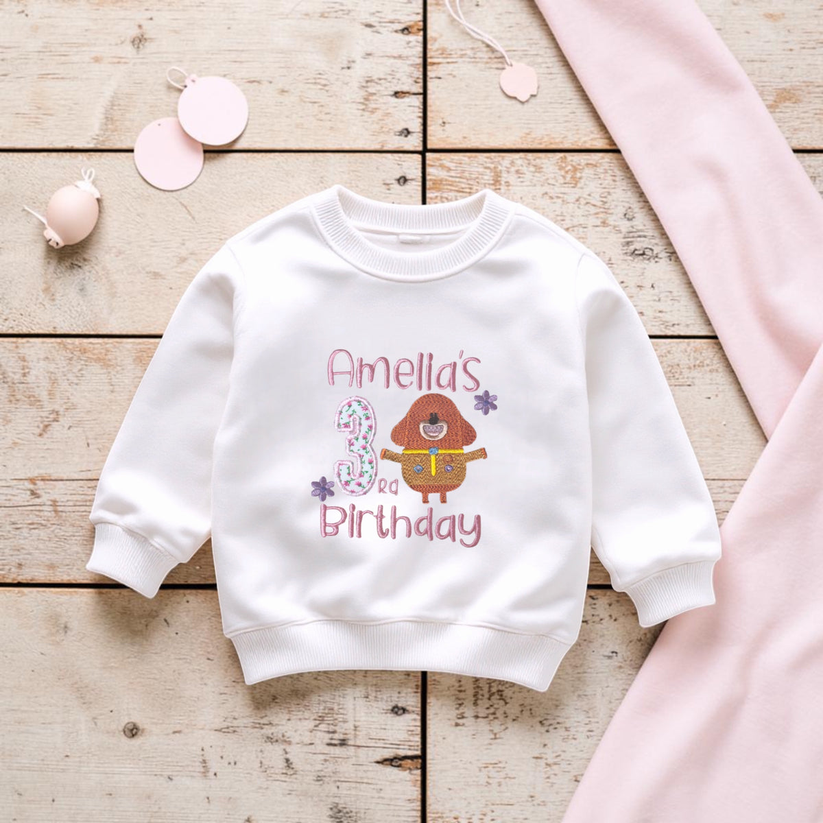 Personalised white girls birthday sweatshirt with embroidered Hey Duggee design, pink name Amelia, floral fabric appliqué number 3 and purple flower details.