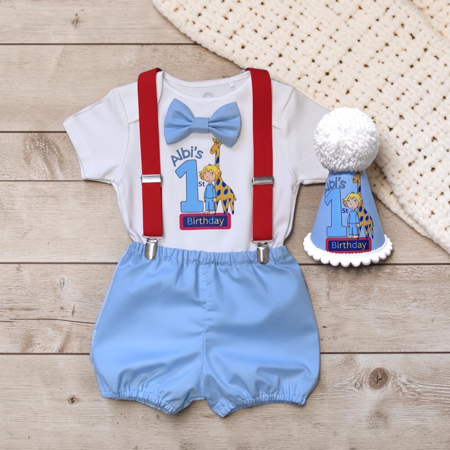 A baby boy's first birthday outfit set laid flat on a light wood background, including a white short-sleeve romper with red suspenders, a blue bow tie, and a blue shorts section with a giraffe and number 1 graphic. A matching blue party hat with a white pom-pom is placed beside it.