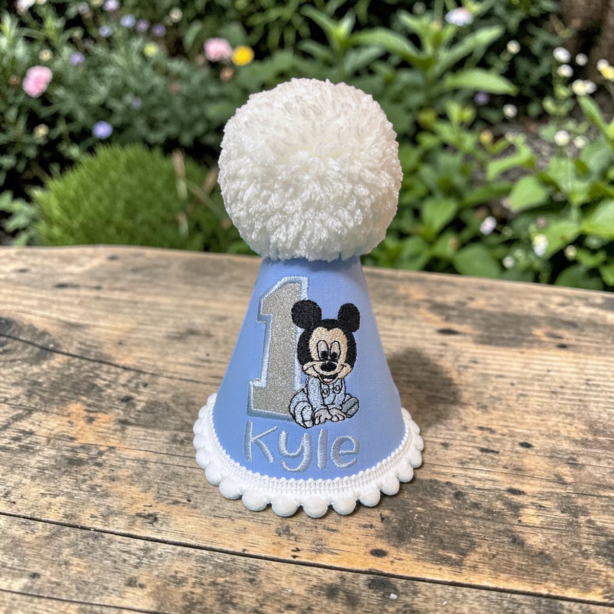 A custom Mickey Mouse first birthday party hat in light blue, featuring a white pom-pom on top, a scalloped white trim, a silver glitter number 1, and a custom name 'Kyle' with a Mickey Mouse illustration. The hat is displayed on a rustic wooden table with a blurred garden background.