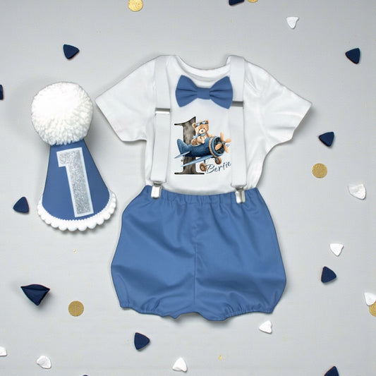 Flat lay of personalised blue aeroplane teddy birthday outfit with printed top, bloomers, braces and handmade party hat.