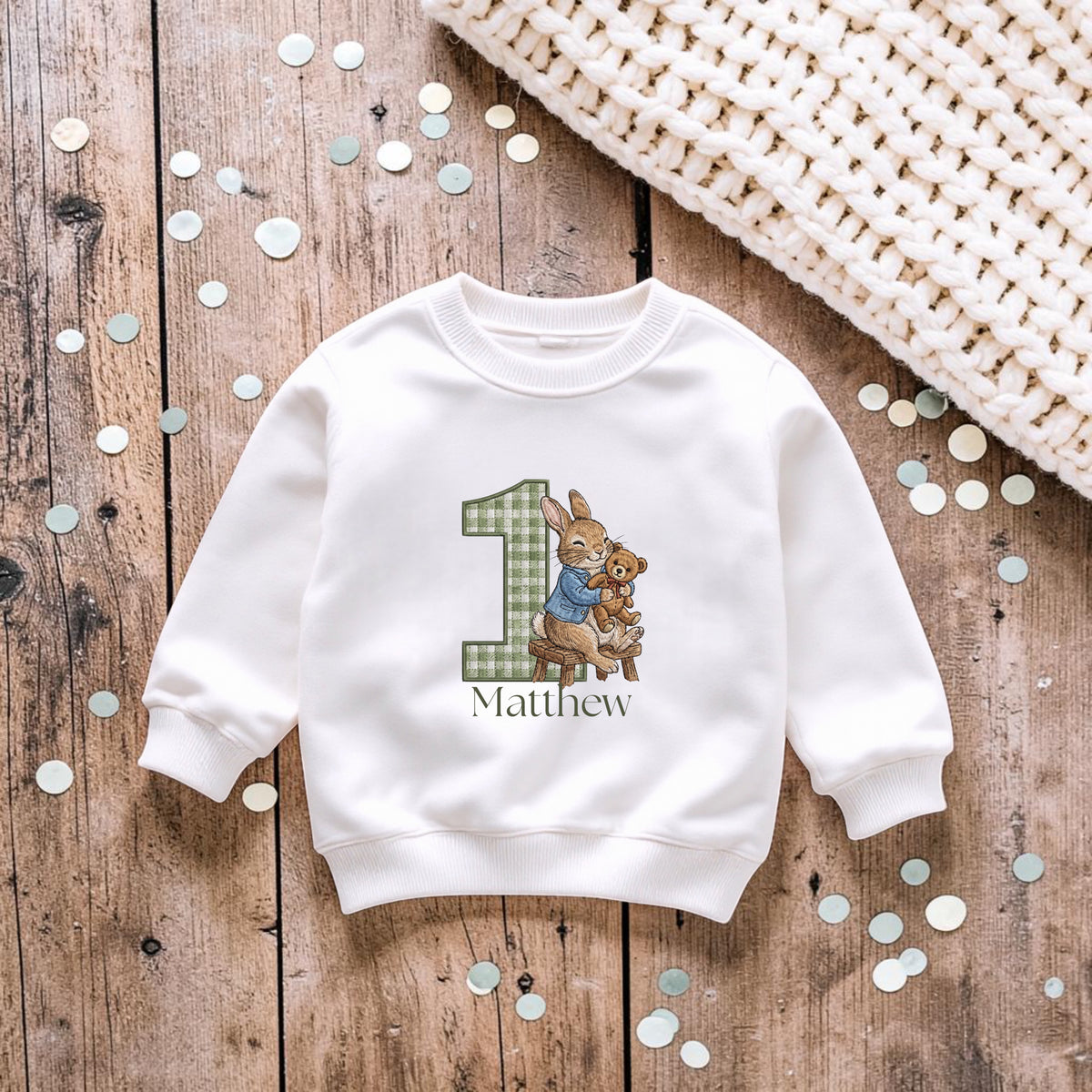 A white long-sleeve sweatshirt laid flat on a rustic wooden surface, featuring a personalized graphic with the number 1 in green plaid, a bunny character in a blue shirt, the name Matthew, and small scattered confetti. A cream knit blanket corner is visible in the top right.