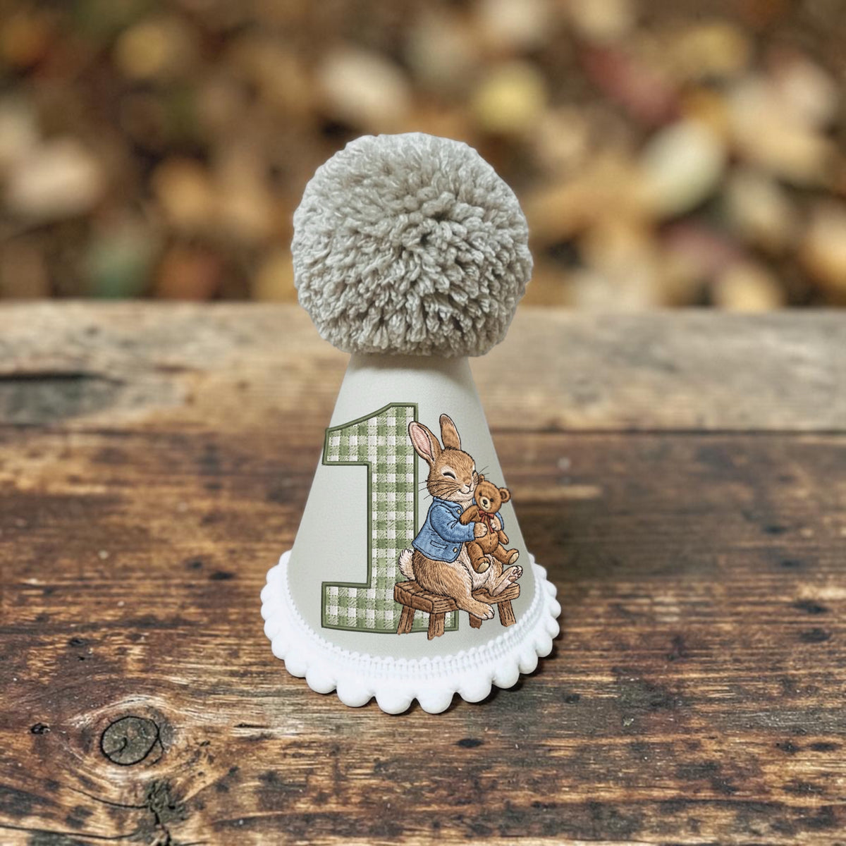 A small white party hat with a gray pom-pom on top, decorated with a green plaid number 1 and an illustration of Beatrix Potter's Peter Rabbit holding a teddy bear. The hat is displayed on a rustic wooden surface with a blurred background of autumn leaves.