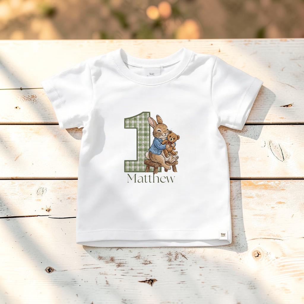 A white short-sleeve baby t-shirt laid flat on a rustic white wooden surface. The shirt features a personalized Easter-themed graphic for a first birthday, showing a large number '1' in a green plaid pattern, a bunny holding a chick, and the name 'Matthew' printed below.