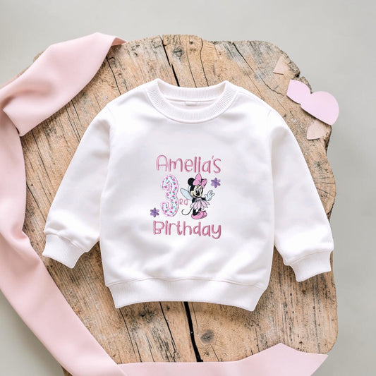 A white long-sleeve sweatshirt for a child, featuring a personalized 'Amelia's 3rd Birthday' graphic with Minnie Mouse. The sweatshirt is displayed on a rustic wooden slice with a pink ribbon and a small pink heart on a light gray background.
