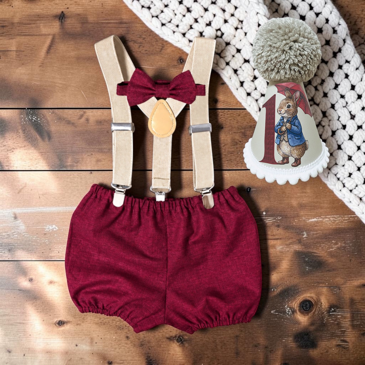 Flat lay of burgundy baby boy cake smash bloomers, beige braces, matching bow and handmade Peter Rabbit birthday hat with umbrella illustration and printed age number.