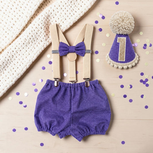 Handmade violet first birthday cake smash set with textured 100% cotton bloomer shorts, matching purple bow, beige suspender braces and coordinating purple party hat with champagne gold number one and fluffy cream pom-pom.