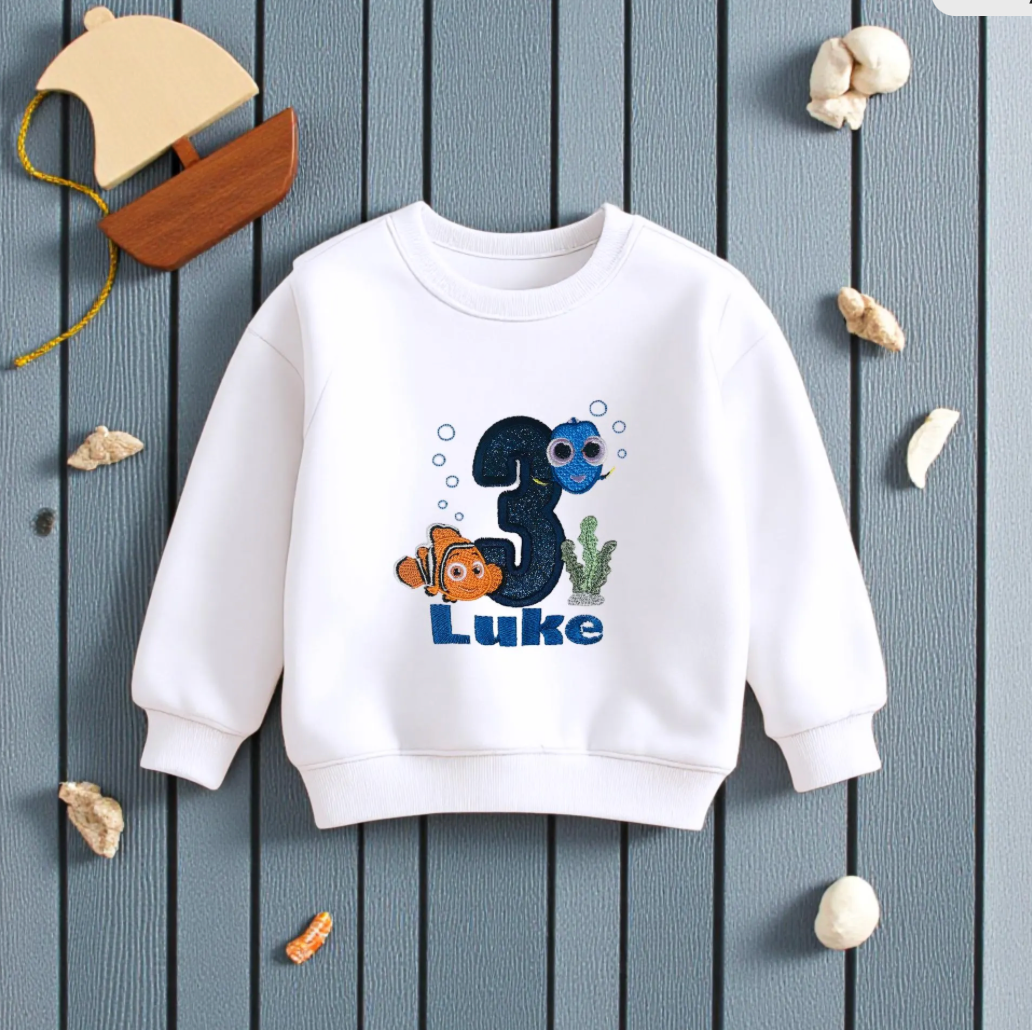 White children's sweatshirt with '3 Luke' design on a wooden surface with decorative items.