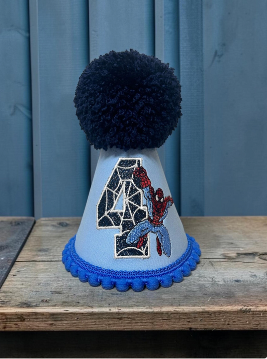 A custom light blue party hat with a dark blue pom-pom, a blue pom-pom trim, and a large embroidered number 4 featuring Spider-Man. The hat is displayed on a rustic wooden table against a blue paneled background.
