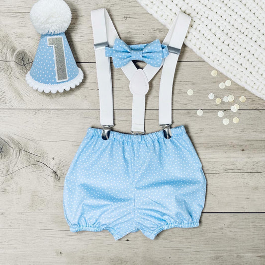 A baby boy's first birthday outfit set featuring light blue and white polka dot bloomers with white suspenders, a matching blue polka dot bow tie, and a blue polka dot party hat with a white pom-pom and silver glitter number 1, arranged on a light wood background.