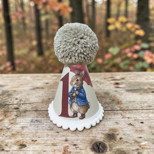 A novelty first birthday party hat featuring a white base with a scalloped edge, a large red number 1, an illustration of Peter Rabbit, and a fluffy stone pom-pom on top. The hat is displayed on a rustic wooden table with a blurred autumn forest background.