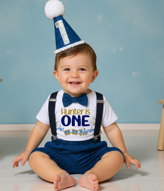 Baby boy wearing personalised blue train first birthday outfit with embroidered bodysuit, suspenders, bow tie and matching blue number one party hat.