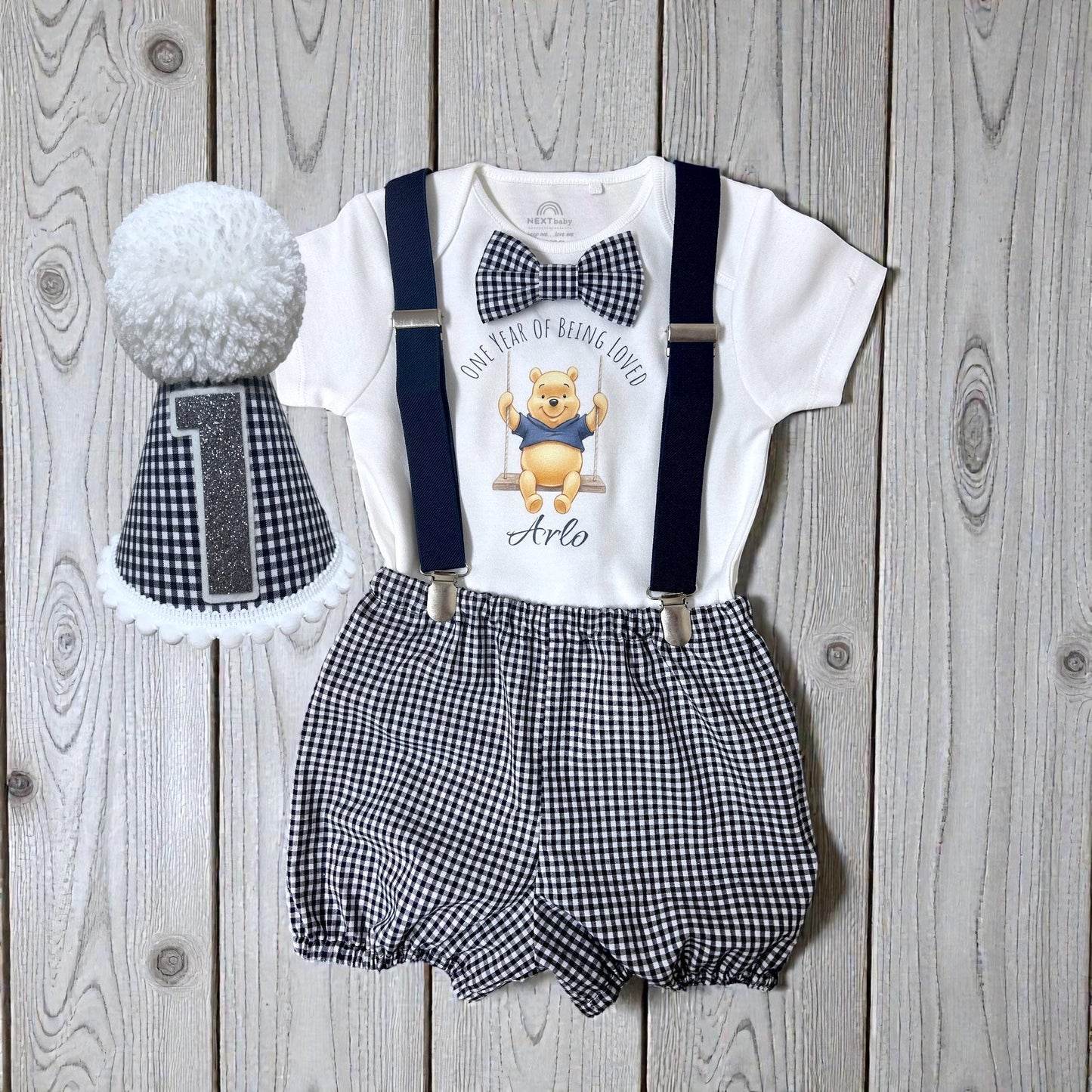 Baby boy cake smash outfit with honey bear printed top, gingham shorts, suspenders and matching party hat styled together on wooden background.