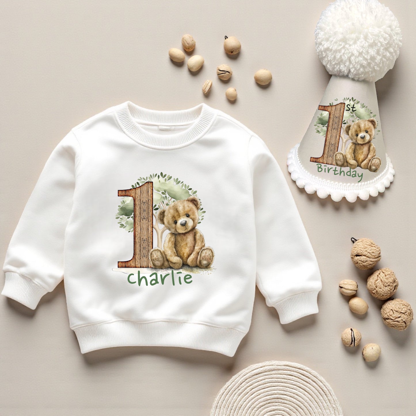 White sweatshirt with teddy bear design and '1st Birthday' text, surrounded by decorative items on a beige background.
