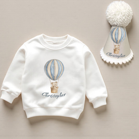Personalised hot air balloon teddy first birthday sweatshirt with matching handmade party hat in soft pastel blue, custom name design.”