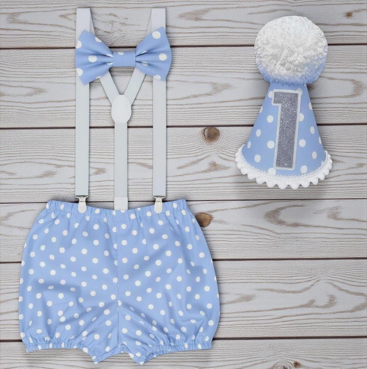 A pair of blue polka dot shorts, a blue bow tie, and a blue birthday hat with the number 1 on it, all laid out on a wooden surface.