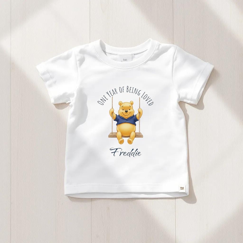 Personalised baby boy birthday T-shirt with honey bear illustration and name, displayed flat on wooden background.