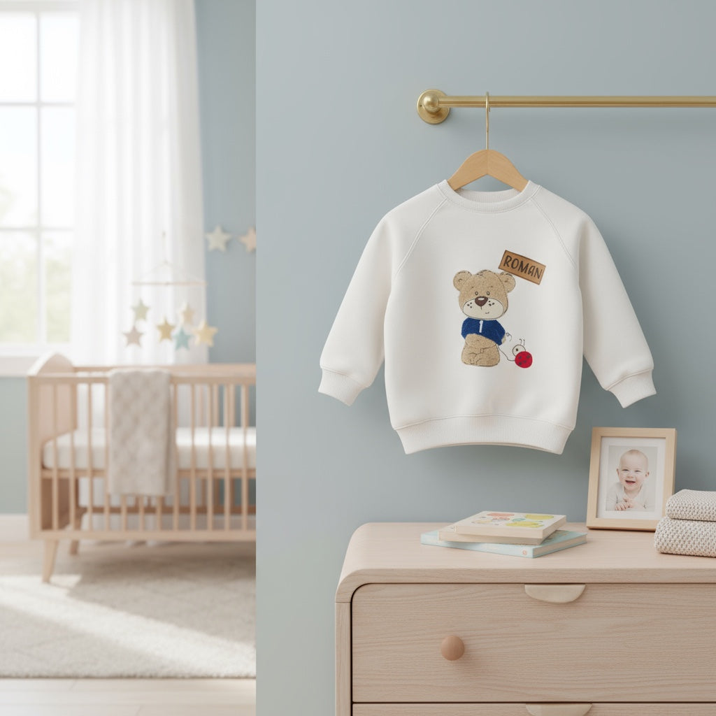 White children's sweatshirt with teddy bear design hanging on a hanger in a nursery.