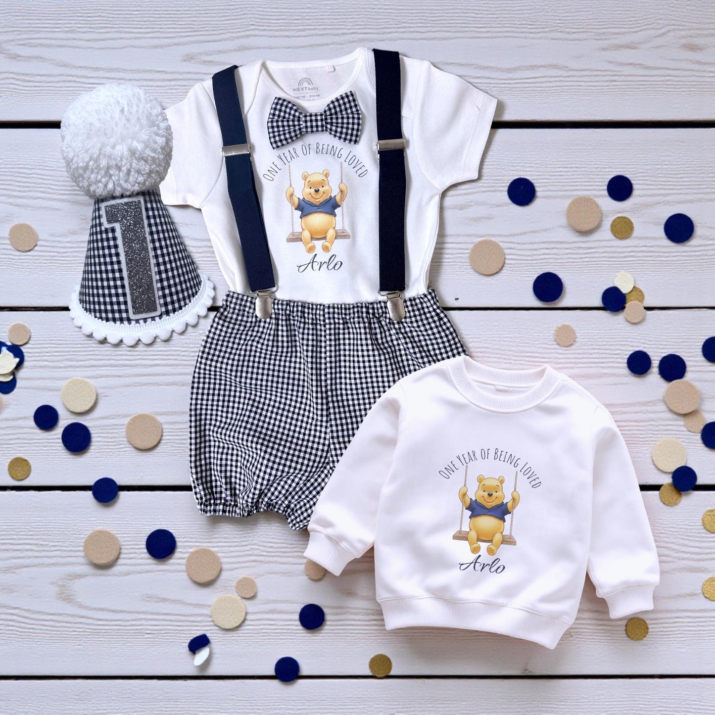 Personalised baby boy birthday outfit with honey bear print, gingham shorts, suspenders, bow tie and matching pom-pom party hat on wooden background.