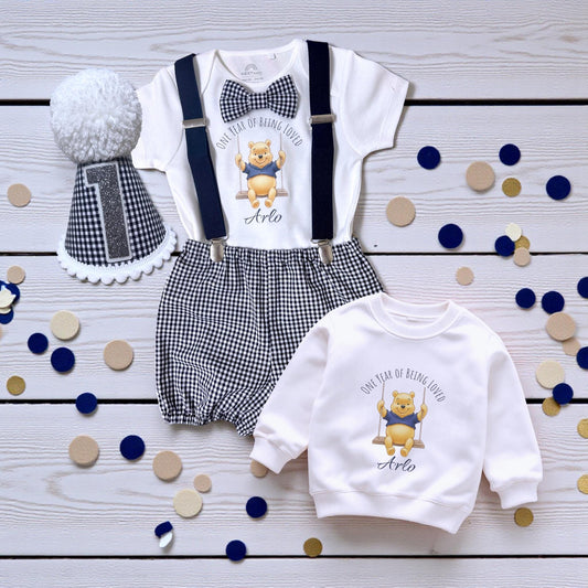 Personalised baby boy birthday outfit with honey bear print, gingham shorts, suspenders, bow tie and matching pom-pom party hat on wooden background.