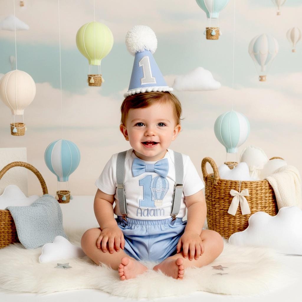 Baby boy wearing blue hot air balloon 1st birthday outfit with suspenders and bow tie, smiling while sitting in nursery, cake smash photo prop for first birthday