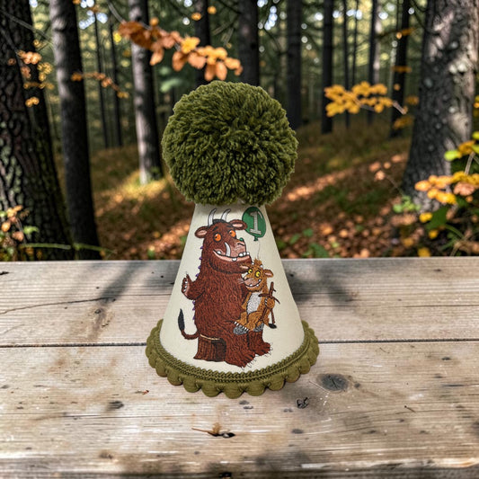 A novelty party hat featuring an illustration of the character Gruffalo topped with a large green pom-pom and a green pompom trim, displayed on a wooden table with a blurred forest background.