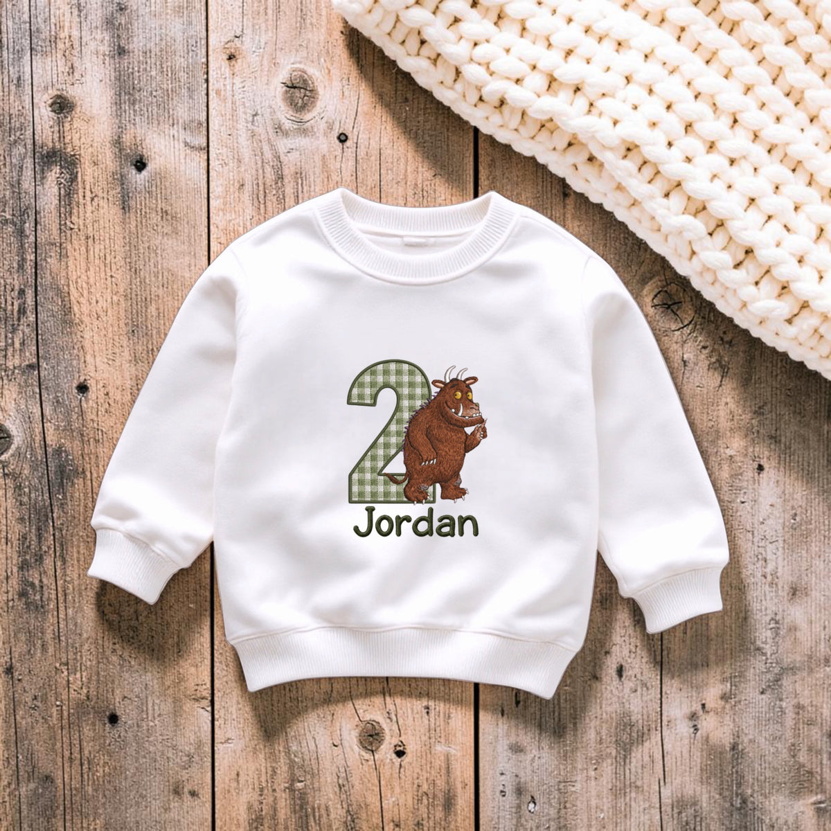 A white long-sleeve sweatshirt for a child, featuring a graphic of a brown monster next to a green plaid number 2, with the name 'Jordan' printed below. The sweatshirt is laid flat on a rustic wooden background next to a chunky cream knit blanket.