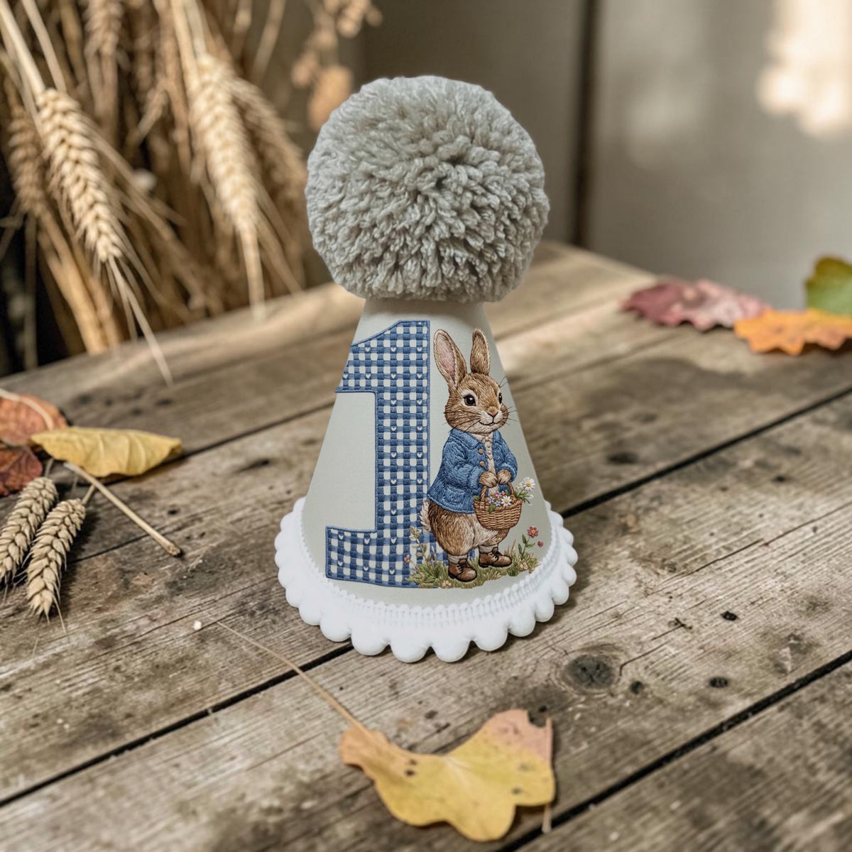 A novelty first birthday hat with a stone pom-pom on top, decorated with a Peter Rabbit illustration and the number 1. The hat is displayed on a rustic wooden table with scattered autumn leaves and dried wheat stalks in the background.