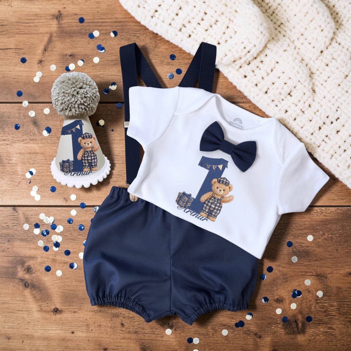 A baby boy's first birthday romper set featuring a white short-sleeve top with a navy bow tie and plaid '1' design, navy blue shorts, and navy suspenders, styled on a wooden surface with a matching party hat and confetti.