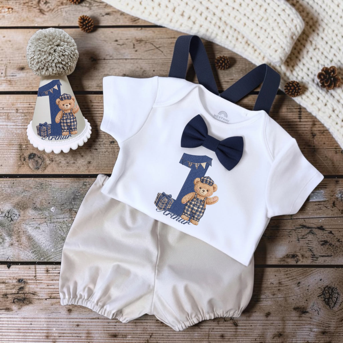 A baby boy's one-piece romper set styled in a flat lay on a rustic wooden surface. The set includes a white short-sleeve romper with navy blue suspenders, a navy blue bow tie, and a graphic of a teddy bear wearing a cap next to a number 1.
