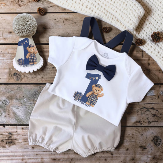 A baby boy's one-piece romper set styled in a flat lay on a rustic wooden surface. The set includes a white short-sleeve romper with navy blue suspenders, a navy blue bow tie, and a graphic of a teddy bear wearing a cap next to a number 1.