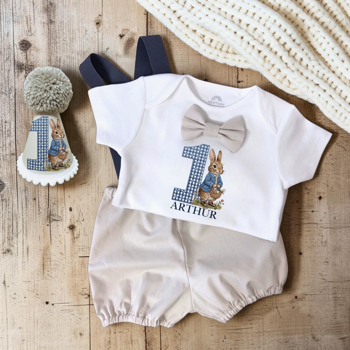 A baby's first birthday outfit laid flat on a rustic wooden surface, featuring a white short-sleeve romper with a gray bow tie, a large blue-and-white gingham number 1 and Peter Rabbit graphic, the name 'ARTHUR' printed below, and cream bloomers. A matching hat with a pom-pom is also shown beside the outfit.