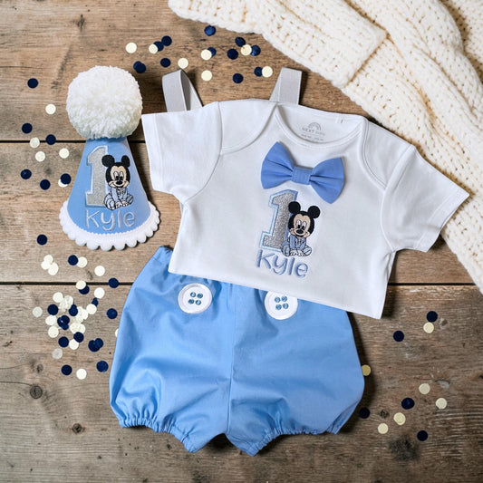 Personalised baby blue Baby Mickey first birthday hat with silver embroidered number one, smiling baby mouse design, name Kyle and oversized white yarn pom-pom.