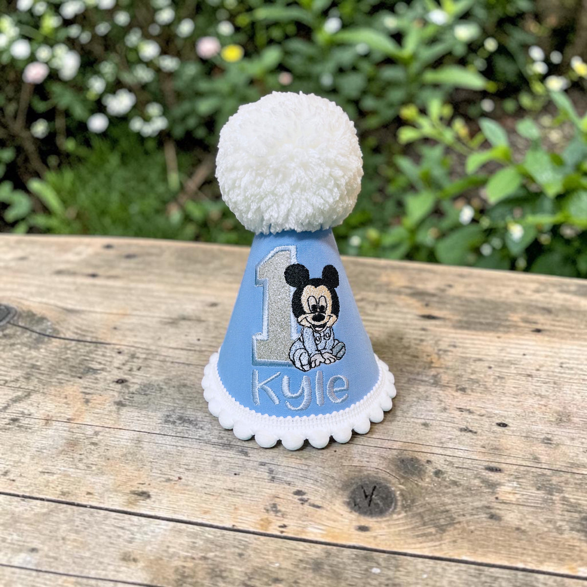 A light blue first birthday party hat with a white pom-pom, personalized with the name 'Kyle' and a Mickey Mouse graphic, sitting on a wooden table with a blurred garden background.