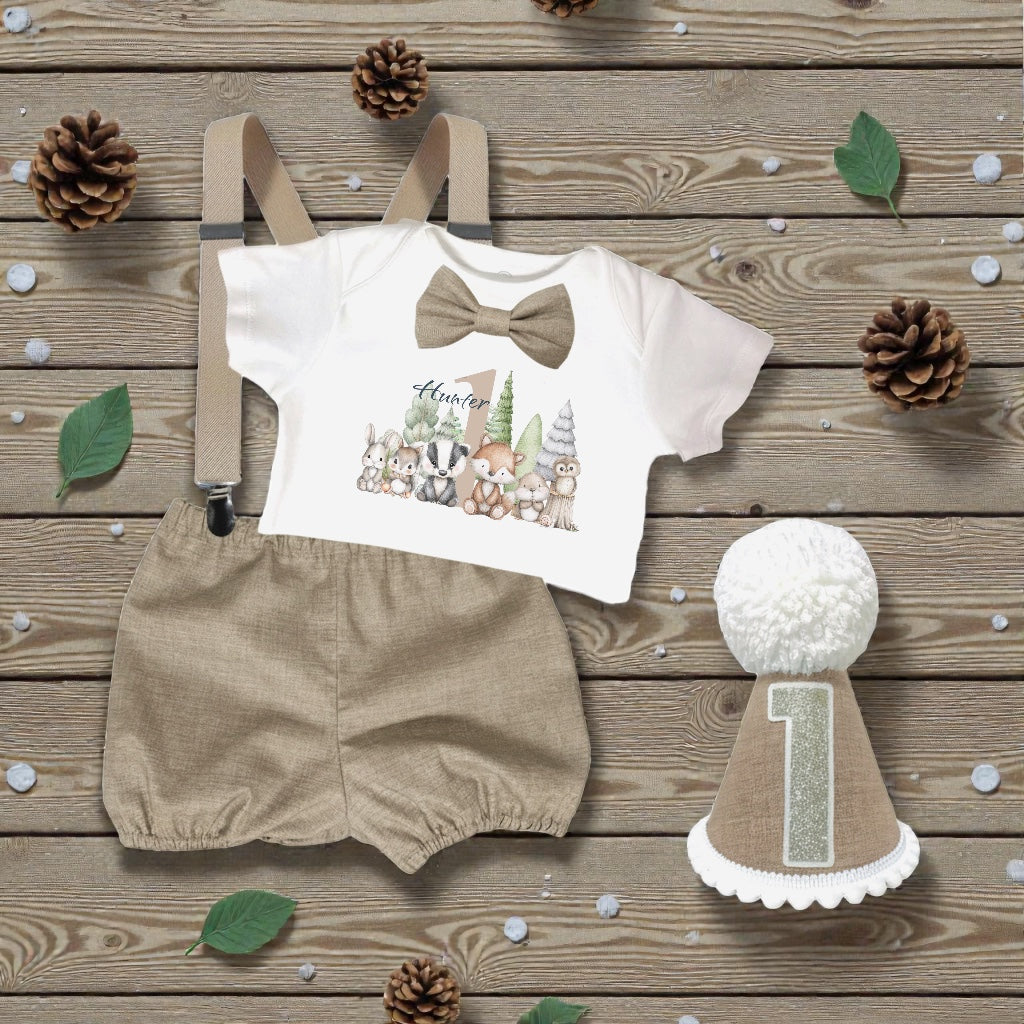 Woodland first birthday outfit flat lay featuring personalised printed bodysuit with forest animals, taupe linen-look bloomers, braces, matching bow tie and handmade taupe birthday hat with number one.