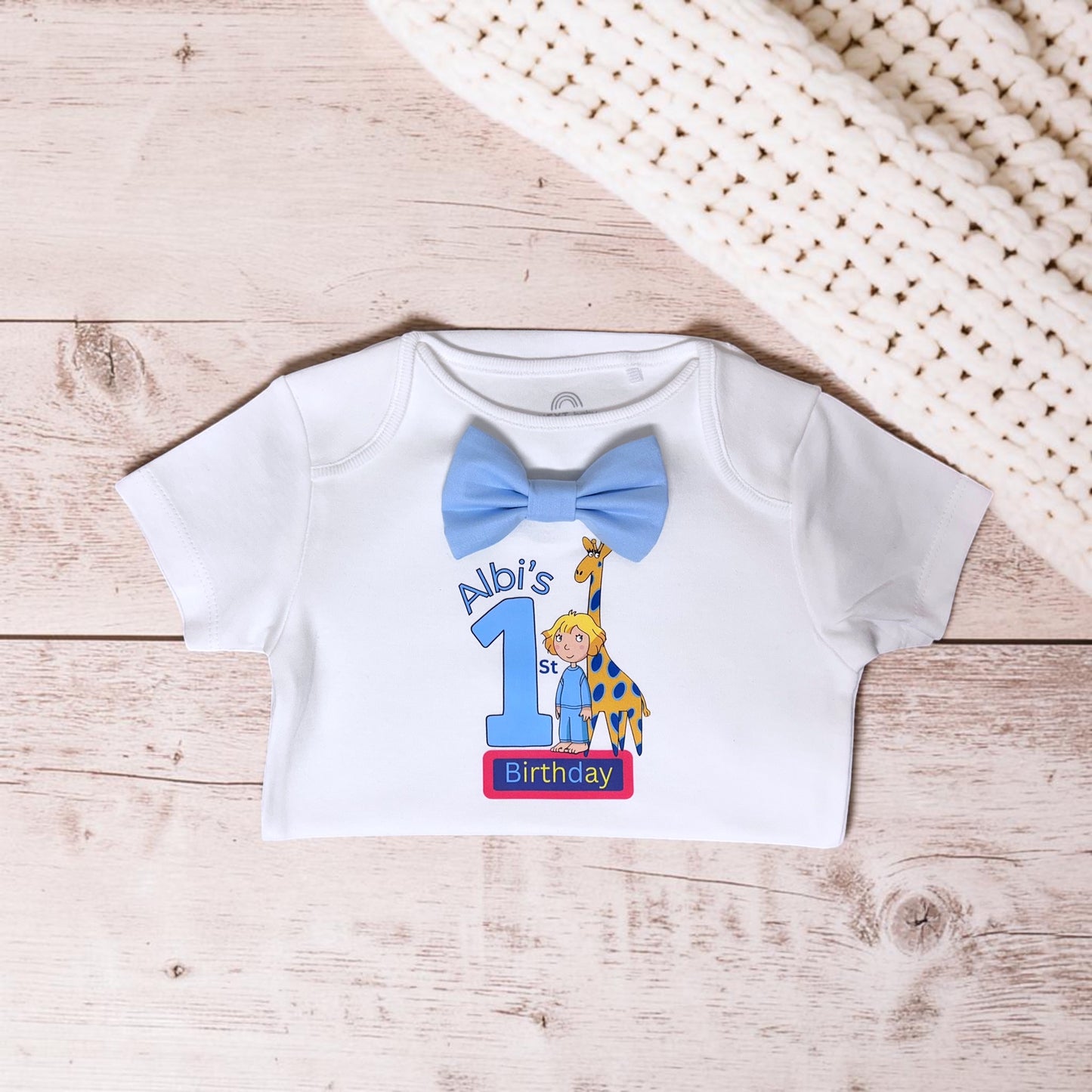 A white short-sleeve baby t-shirt laid flat on a light wood surface, featuring a custom birthday graphic with a blue bow tie, the number 1, a cartoon boy, a giraffe, and the text 'Abi's 1st Birthday'. A cream-colored knit blanket is partially visible in the top right corner.