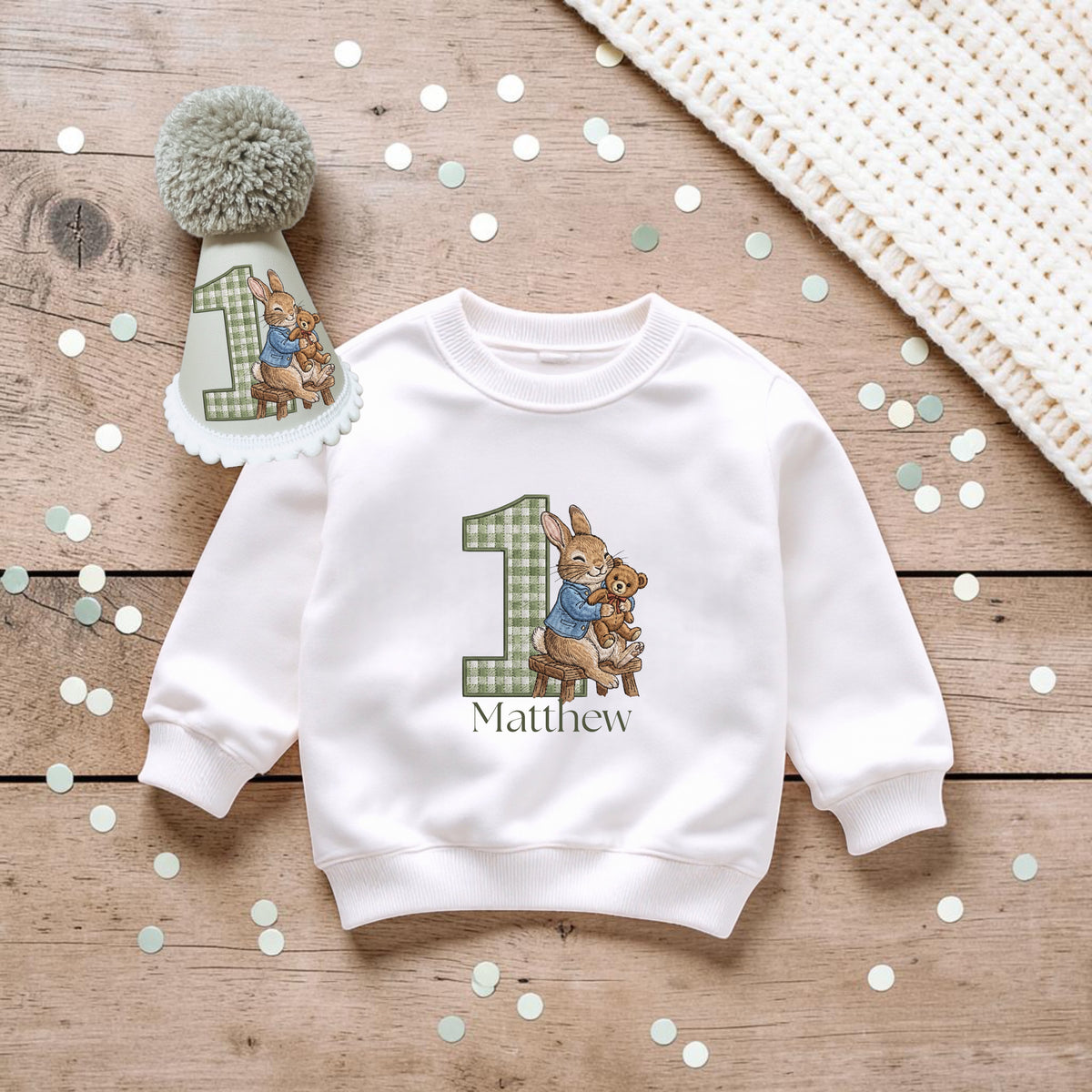 A white long-sleeve sweatshirt for a baby's first birthday, featuring a graphic of Beatrix Potter's rabbit characters inside a green gingham number 1, with the name 'Matthew' printed below. The sweatshirt is styled on a wooden surface with a matching first birthday hat and confetti.