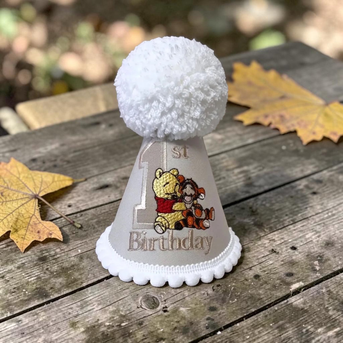 A white first birthday party hat with a fluffy white pom-pom on top, featuring Winnie the Pooh and Tigger graphics with the text '1st Birthday' on a weathered wooden table with autumn leaves.