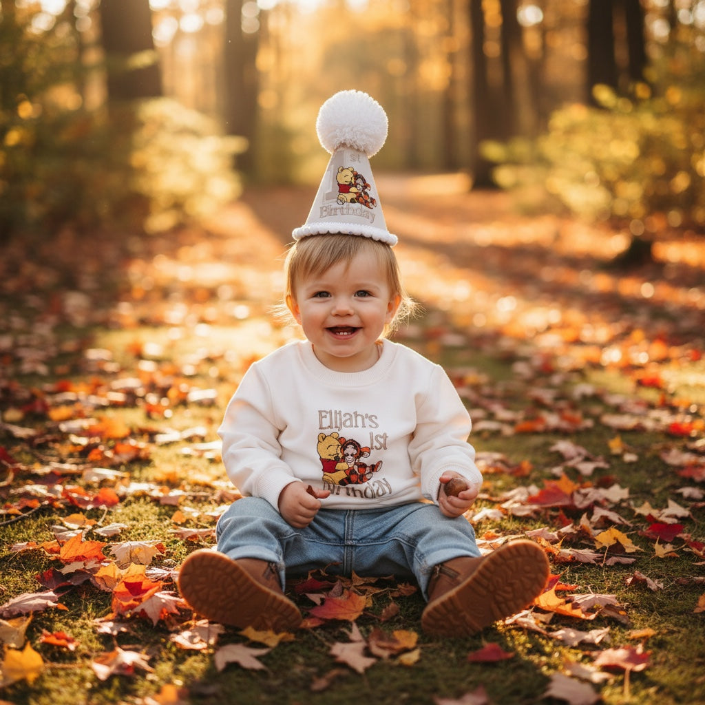 A baby sitting outdoors on a leaf-covered grassy path, wearing a white long-sleeve shirt with a cartoon graphic, blue jeans, brown boots, and a white party hat with a pom-pom.