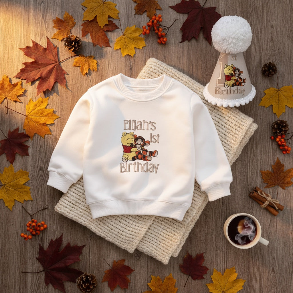 A white crewneck sweatshirt with a 'Ellia's 1st Birthday' cartoon graphic, styled in a flat lay on a wooden surface with autumn leaves, a knit blanket, a party hat, and a cup of coffee.